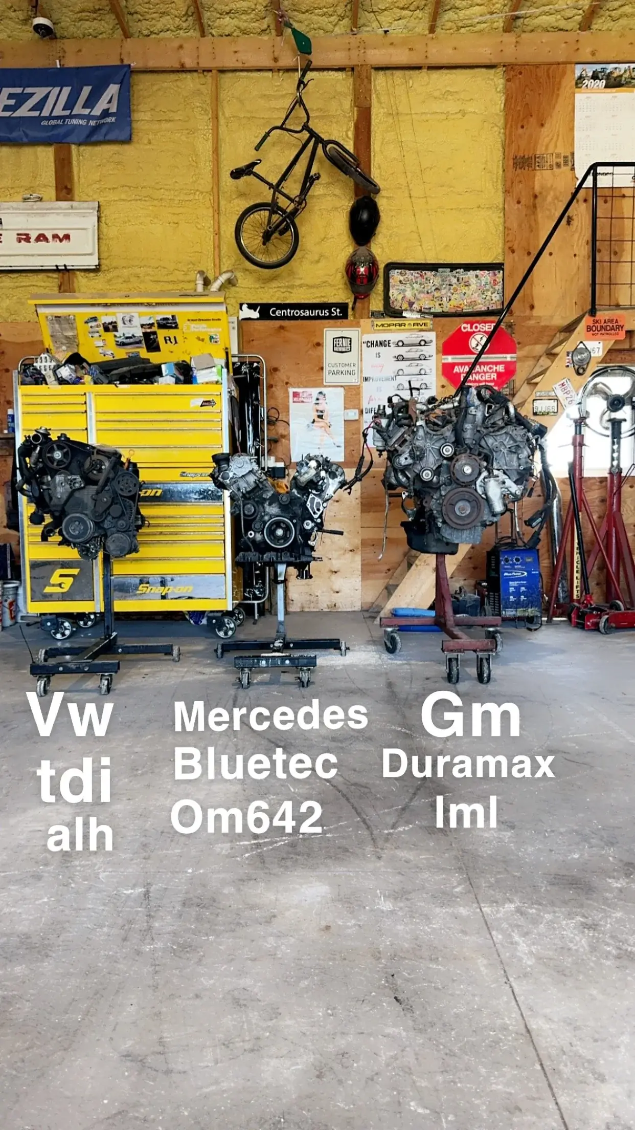 Garage with three cars' engines on engine stands, a yellow tool chest, a wall with a bicycle, helmets, and various signs including street names and warning signs.